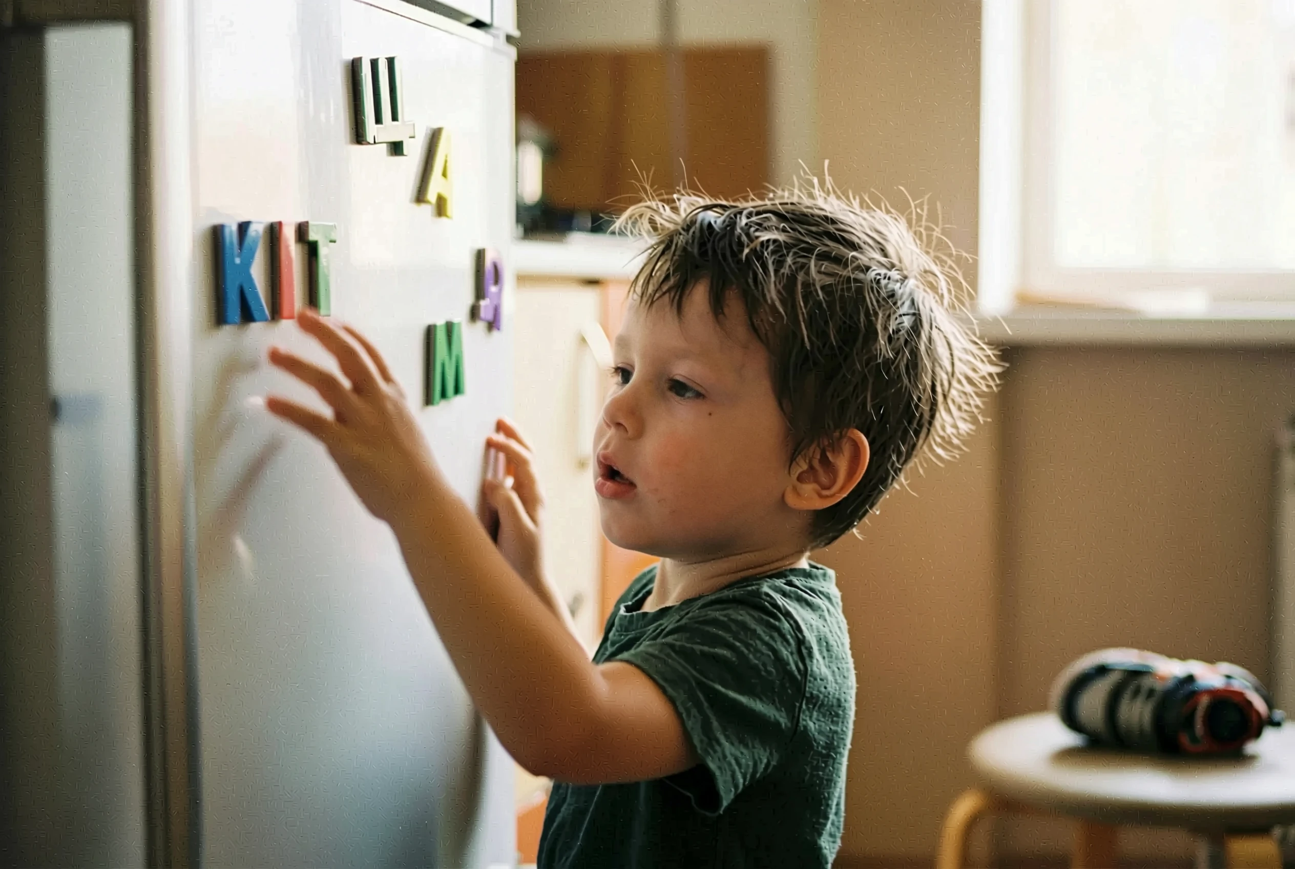 Хлопчик стоїть біля холодильника і складає слово з магнітних літер: на дверях холодильника видно кириличні літери-магніти, дитина тягнеться замінити одну літеру
