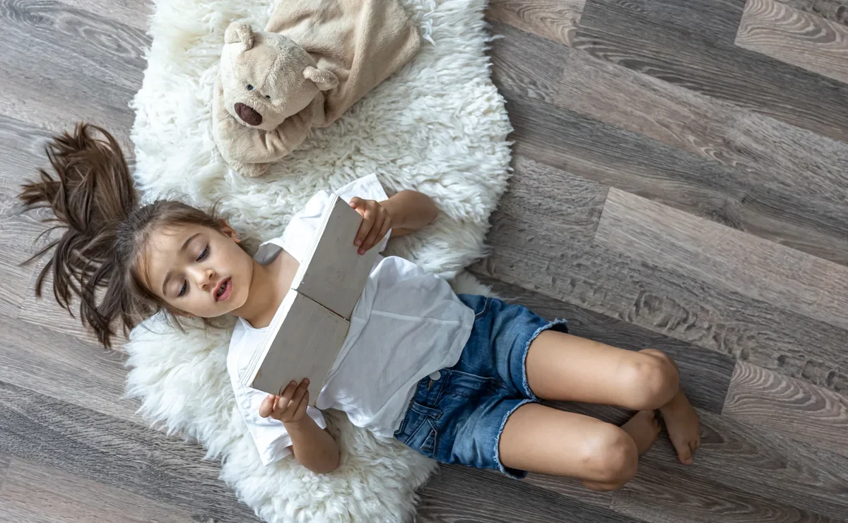 Дитина дошкільного віку розглядає ілюстрацію в книжці, лежачи на підлозі